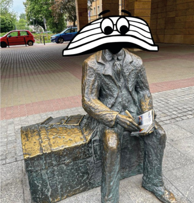 A bronze statue of a man in a suit sitting on a bench and reading a newspaper, photographed outdoors on a paved street. The statue's face area is obscured by a cartoon-style black-and-white hat graphic.